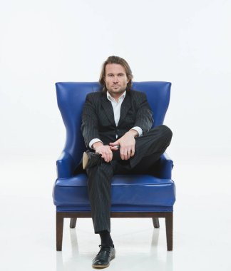 Siyah takım elbiseli yakışıklı adam mavi bir sandalyede oturuyor, üzgün, uzun saçlı, kaba adam, beyaz gömlek, iş adamı, çok şık, kameraya bakıyor.