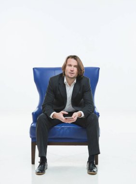 Siyah takım elbiseli yakışıklı adam mavi bir sandalyede oturuyor, üzgün, uzun saçlı, kaba adam, beyaz gömlek, iş adamı, çok şık, kameraya bakıyor.
