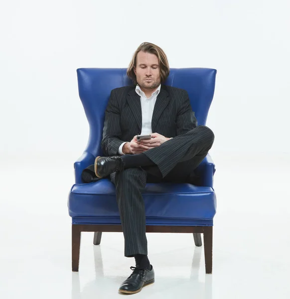 Os homens bonitos de terno preto se senta em uma cadeira azul, ele é triste, homem brutal com cabelos longos, camisa branca, homem de negócios, muito elegante, olhando no smartphone — Fotografia de Stock