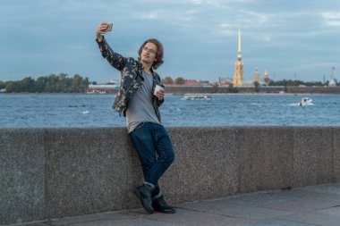 Yakışıklı adamlar birini bekliyor, kahve içiyor ve telefona bakıyor, asker ceketi ve kot pantolon giyiyor, Peter ve Paul kalesi arka planda.