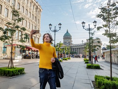 Yakışıklı adam selfie çekiyor. Dışarıdan bak, sarı bir kazak giyiyor, siyah bir yağmurluk ya da ceket onun elleri, Bolshaya Konyushennaya sokağı ve arka planda Kazan Katedrali, güneşli bir gün.