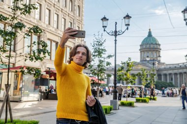 Yakışıklı adam selfie çekiyor. Dışarıdan bak, sarı bir kazak giyiyor, siyah bir yağmurluk ya da ceket onun elleri, Bolshaya Konyushennaya sokağı ve arka planda Kazan Katedrali, güneşli bir gün.