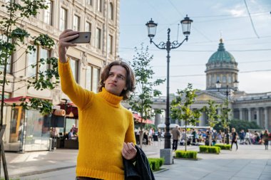 Yakışıklı adam selfie çekiyor. Dışarıdan bak, sarı bir kazak giyiyor, siyah bir yağmurluk ya da ceket onun elleri, Bolshaya Konyushennaya sokağı ve arka planda Kazan Katedrali, güneşli bir gün.