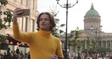 Yakışıklı adam selfie çekiyor. Dışarıdan bak, sarı bir kazak giyiyor, siyah bir yağmurluk ya da ceket onun elleri, Bolshaya Konyushennaya sokağı ve arka planda Kazan Katedrali, güneşli bir gün.