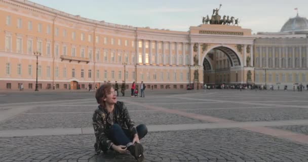 Красивий молодий чоловік, що сидить на площі палацу і п'є каву на заході сонця, одягнений у військовий піджак і джинси, довгі кучеряві волоски, Арка Генерального штабу будівля на задньому плані — стокове відео