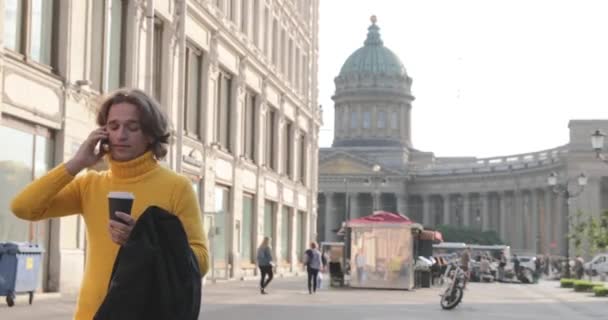 Pria tampan sedang menunggu seseorang, minum kopi, ia mengenakan sweater kuning dan jas hujan hitam, Bolshaya Konyushennaya jalan dan Kazan Katedral di latar belakang, hari yang cerah — Stok Video