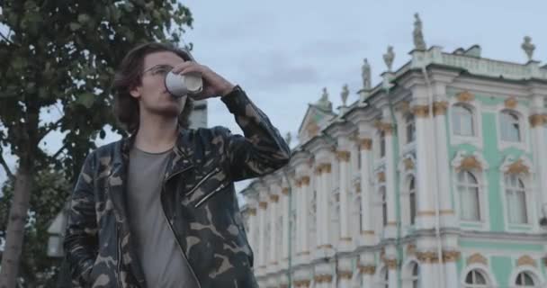 The handsome men is waiting someone, drinks coffee and looks in phone, he is dressed in a military jacket and jeans, State Hermitage museum is on background — Stock Video