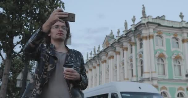 Les beaux hommes attendent quelqu'un, boit du café et regarde au téléphone, il est vêtu d'une veste militaire et d'un jean, le musée de l'Ermitage est en arrière-plan — Video