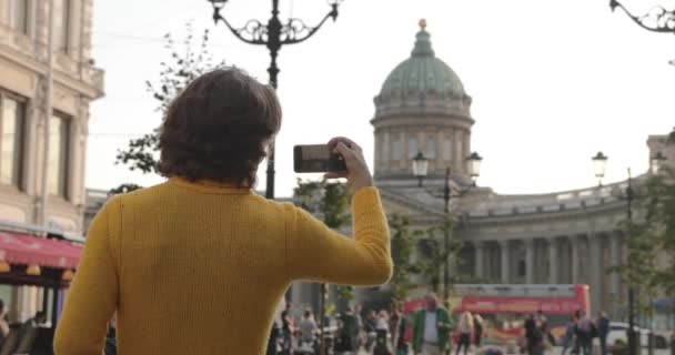 Przystojny mężczyzna robi selfie, Spójrz z zewnątrz, ubrany w żółty sweter, czarny płaszcz przeciwdeszczowy lub kurtka jest jego ręce, Bolshaya Konyushennaya ulicy i Kazan Cathedral na tle, słoneczny dzień — Wideo stockowe