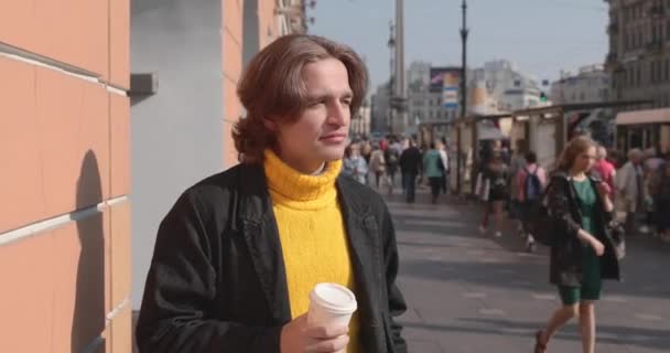 Ο όμορφος άντρας περιμένει κάποιον, πίνει καφέ, ντύνεται με ένα κίτρινο πουλόβερ και μαύρο αδιάβροχο, δρόμο της Αγίας Πετρούπολης και πολλοί πεζοί είναι στο παρασκήνιο, ηλιόλουστη μέρα — Αρχείο Βίντεο