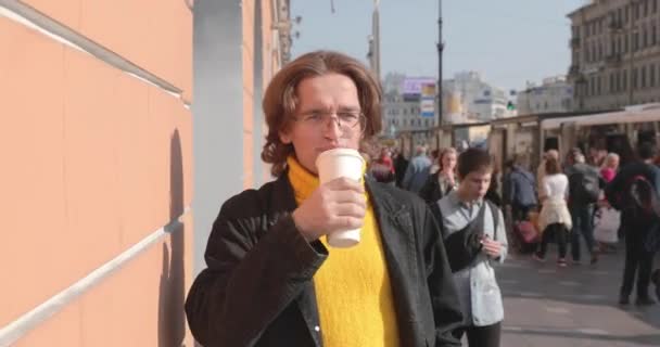 Красивые мужчины ждут кого-то, пьют кофе, он одет в желтый свитер и черный плащ, улица Санкт-Петербурга и много пешеходов на заднем плане, солнечный день — стоковое видео