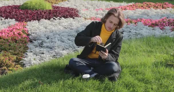 Den unge mannen dricker kaffe och läser boken i parken, han ler, vilar, är klädd i en gul tröja, blommor och gräs på bakgrunden, solig dag — Stockvideo