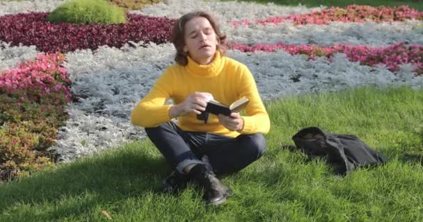 Молодой человек пьет кофе и читает книгу в парке, улыбается, отдыхает, одет в желтый свитер, цветы и траву на заднем плане, солнечный день — стоковое видео