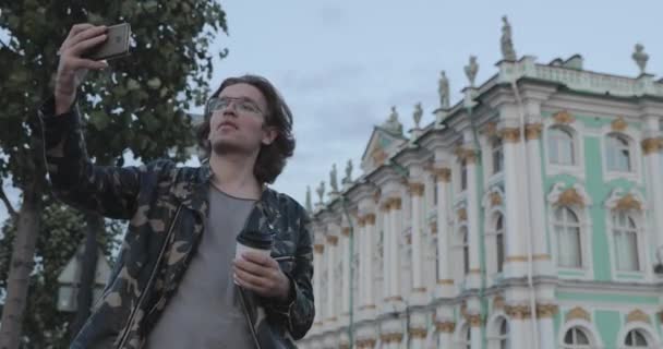 Les beaux hommes attendent quelqu'un, boit du café et regarde au téléphone, il est vêtu d'une veste militaire et d'un jean, le musée de l'Ermitage est en arrière-plan — Video