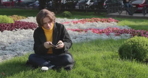 The young man drinks coffee and reads the book in the park, he smiles, has a rest, is dressed in a yellow sweater, flowers and grass on background, sunny day — Stock Video