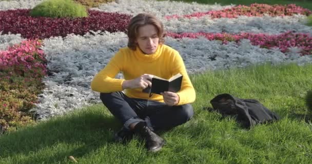 De jongeman drinkt koffie en leest het boek in het park, hij glimlacht, heeft rust, is gekleed in een gele trui, bloemen en gras op de achtergrond, zonnige dag — Stockvideo