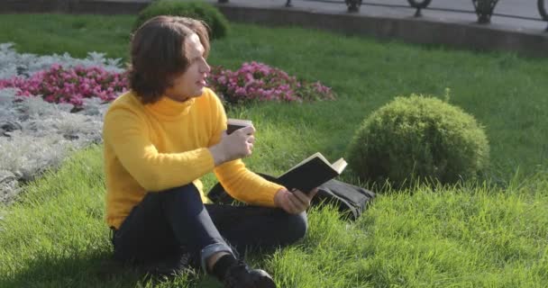 El joven bebe café y lee el libro en el parque, sonríe, descansa, se viste con un suéter amarillo, flores y hierba en el fondo, día soleado — Vídeos de Stock