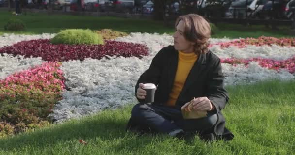 Tânărul bea cafea și citește cartea în parc, zâmbește, se odihnește, este îmbrăcat într-un pulover galben, flori și iarbă pe fundal, zi însorită — Videoclip de stoc