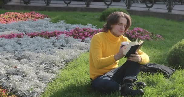 The young man drinks coffee and reads the book in the park, he smiles, has a rest, is dressed in a yellow sweater, flowers and grass on background, sunny day — Stock Video