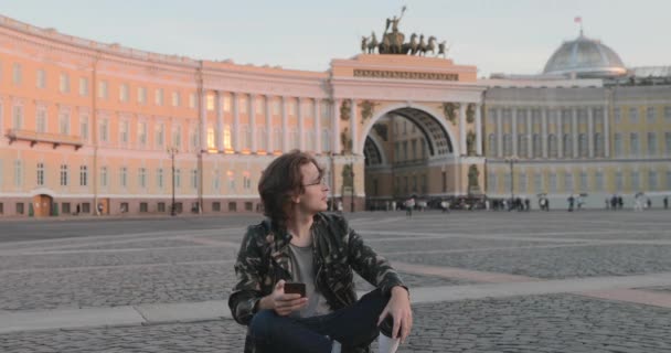 Красивий молодий чоловік, що сидить на площі палацу і п'є каву на заході сонця, одягнений у військовий піджак і джинси, довгі кучеряві волоски, Арка Генерального штабу будівля на задньому плані — стокове відео