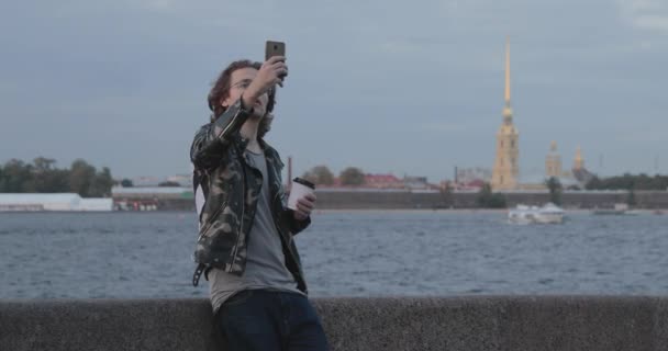 The handsome men is waiting someone, drinks coffee and looks in phone, he is dressed in a military jacket and jeans, Peter and Paul fortress is on background — Stock Video