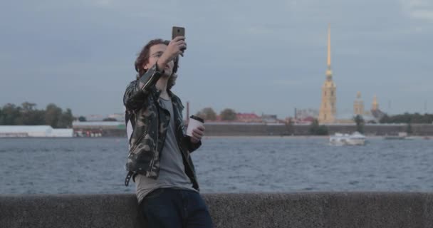 De knappe mannen wachten op iemand, drinken koffie en kijkt in de telefoon, hij is gekleed in een militaire jas en jeans, Peter en Paul fort is op de achtergrond — Stockvideo