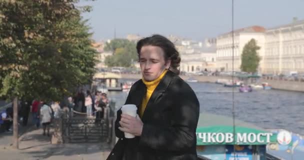 Os homens bonitos está esperando alguém, bebe café, ele vestiu uma camisola amarela e capa de chuva preta, aterro do rio Fontanka e barco está no fundo, um monte de pedestre, dia ensolarado — Vídeo de Stock