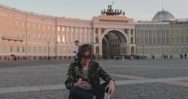 Der schöne junge Mann sitzt auf dem Palastplatz und trinkt bei Sonnenuntergang Kaffee, er trägt eine Militärjacke und Jeans, lange lockige Haare, im Hintergrund ist der Bogen des Generalstabsgebäudes zu sehen — Stockvideo