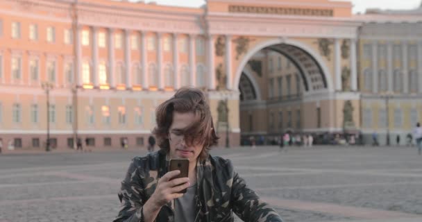 Ο όμορφος νεαρός που κάθεται στην πλατεία του Παλατιού και πίνει καφέ το ηλιοβασίλεμα, ντυμένος με στρατιωτικό μπουφάν και τζιν, μακριά σγουρά μαλλιά, Arch of the General Staff Building is on background — Αρχείο Βίντεο