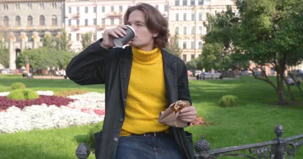 Молодий чоловік п'є каву і їсть солодкий торт в парку, він посміхається, одягнений в жовтий светр і чорний плащ або піджак, джинсові штани, квіти і зелена трава на фоні, сонячний день — стокове відео