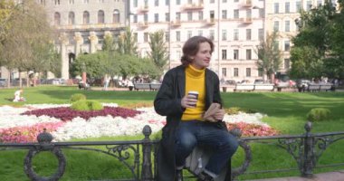 Genç adam kahve içiyor ve parkta tatlı kek yiyor, gülümsüyor, sarı bir kazak ve siyah bir ceket giyiyor, kot pantolon, çiçekler ve arka planda yeşil çimenler, güneşli bir gün.