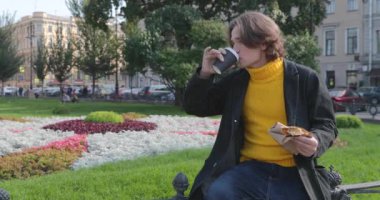 Genç adam kahve içiyor ve parkta tatlı kek yiyor, gülümsüyor, sarı bir kazak ve siyah bir ceket giyiyor, kot pantolon, çiçekler ve arka planda yeşil çimenler, güneşli bir gün.
