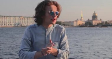 Güneş gözlüklü yakışıklı genç adam gün batımında hareketli bir şey söylüyor, uzun kıvırcık saçlı, kot pantolon giymiş, arka planda St. Petersburg 'un simgeleri var.