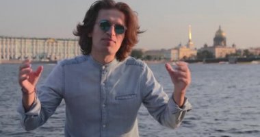 Güneş gözlüklü yakışıklı genç adam gün batımında hareketli bir şey söylüyor, uzun kıvırcık saçlı, kot pantolon giymiş, arka planda St. Petersburg 'un simgeleri var.