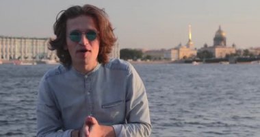 Güneş gözlüklü yakışıklı genç adam gün batımında hareketli bir şey söylüyor, uzun kıvırcık saçlı, kot pantolon giymiş, arka planda St. Petersburg 'un simgeleri var.