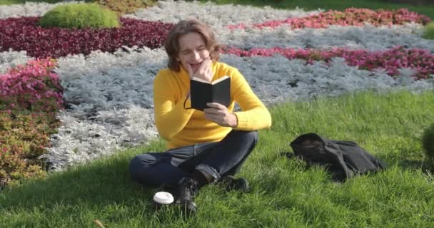 O jovem bebe café e lê o livro no parque, sorri, descansa, veste-se com uma camisola amarela, flores e grama no fundo, dia ensolarado — Vídeo de Stock