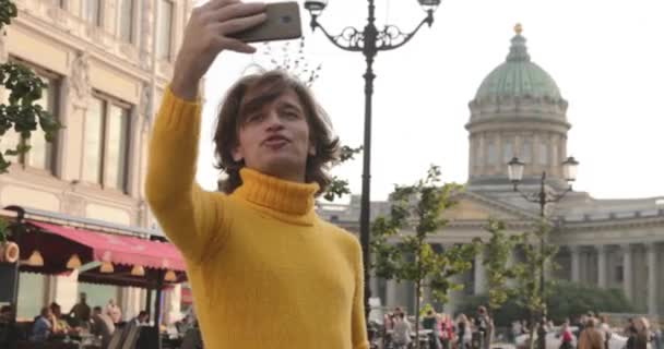 Os homens bonitos faz selfie, Olhe de fora, ele vestiu uma camisola amarela, uma capa de chuva preta ou jaqueta é suas mãos, Bolshaya Konyushennaya rua e Catedral Kazan no fundo, dia ensolarado — Vídeo de Stock