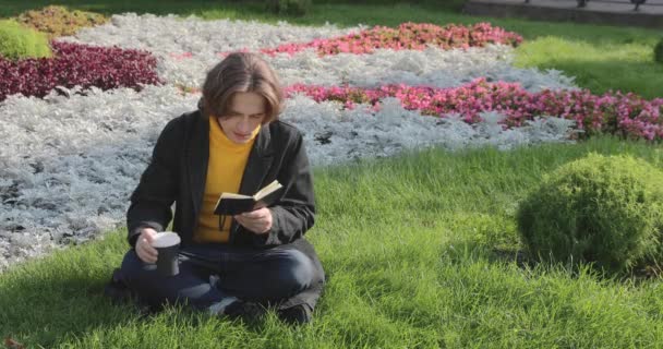 El joven bebe café y lee el libro en el parque, sonríe, descansa, se viste con un suéter amarillo, flores y hierba en el fondo, día soleado — Vídeos de Stock