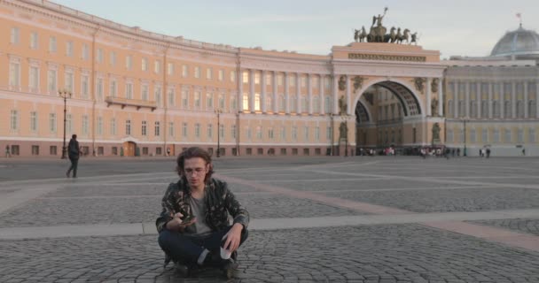 Le beau jeune homme assis sur la place du Palais et boit du café au coucher du soleil, il vêtu d'une veste militaire et d'un jean, de longs poils bouclés, l'Arc de l'état-major général est en arrière-plan — Video