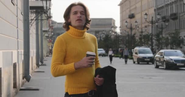 Il bell'uomo è in attesa di qualcuno, beve caffè, si è vestito con un maglione giallo e impermeabile nero, Bolshaya Konyushennaya strada e Kazan Cattedrale sullo sfondo, giornata di sole — Video Stock