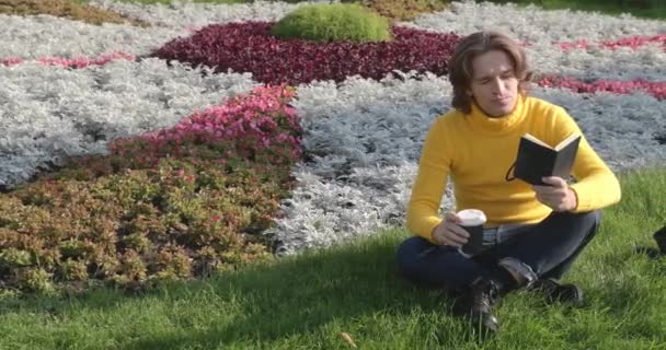 El joven bebe café y lee el libro en el parque, sonríe, descansa, se viste con un suéter amarillo, flores y hierba en el fondo, día soleado — Vídeo de stock
