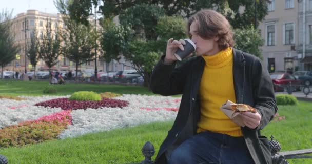 Der junge Mann trinkt Kaffee und isst süßen Kuchen im Park, er lächelt, trägt einen gelben Pullover und schwarze Regenjacke, Jeanshose, Blumen und grünes Gras im Hintergrund, sonniger Tag — Stockvideo