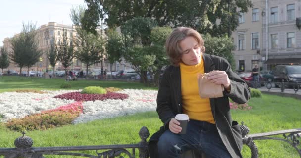 De jongeman drinkt koffie en eet zoete cake in het park, hij glimlacht, is gekleed in een gele trui en zwarte regenjas, Jeans broek, bloemen en groen gras op de achtergrond, zonnige dag — Stockvideo