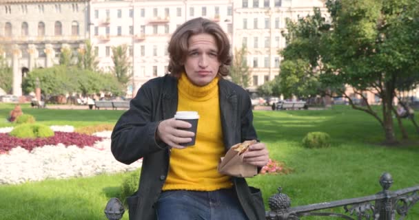 Il giovane beve caffè e mangia dolci dolci nel parco, sorride, è vestito con un maglione giallo e impermeabile nero o giacca, pantaloni Jeans, fiori e erba verde sullo sfondo, giornata di sole — Video Stock
