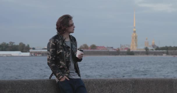 Les beaux hommes attendent quelqu'un, boit du café et regarde au téléphone, il est vêtu d'une veste militaire et un jean, Peter et Paul forteresse est sur le fond — Video