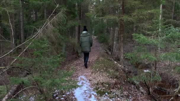 La chica con una chaqueta verde, pantalones negros y una gorra va por un sendero hacia el bosque entre árboles, un paisaje primaveral, silencio, privacidad, caminar al aire libre, La persona nos deja, Aislamiento — Vídeo de stock