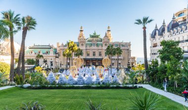 Monaco, Monte-Carlo, 25 Aralık 2019: Günbatımında Monte-Carlo Meydanı, beyaz Noel ağaçları, Paris Oteli, güneşli gün, Noel süsü, turistler, çeşme, yeni evler, yeşil çimenler