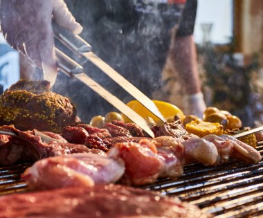 Et ızgarada salamura ediliyor, kaburga yayılıyor, kazık, sosis, kenarlar, tavuk, çıplak alev, karabiber, duman, yakacak odun, şefin elleri, eldivenli eller, yakın çekim.