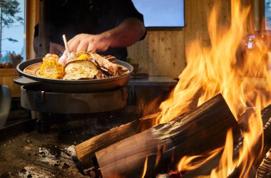 Bir tavada sebzeli büyük tabak, sarımsak, tatlı biber, patates, mısır ve patlıcan, büyük çıplak ateşin yanında, kırmızı kömür, duman, yakacak odun, arka planda beyaz ekran