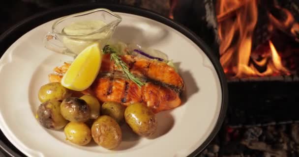 Grillowana ryba jest na białym talerzu z ziemniakami, cytryną i sosem, nagi płomień, czerwone węgle, dym, drewno opałowe, — Wideo stockowe
